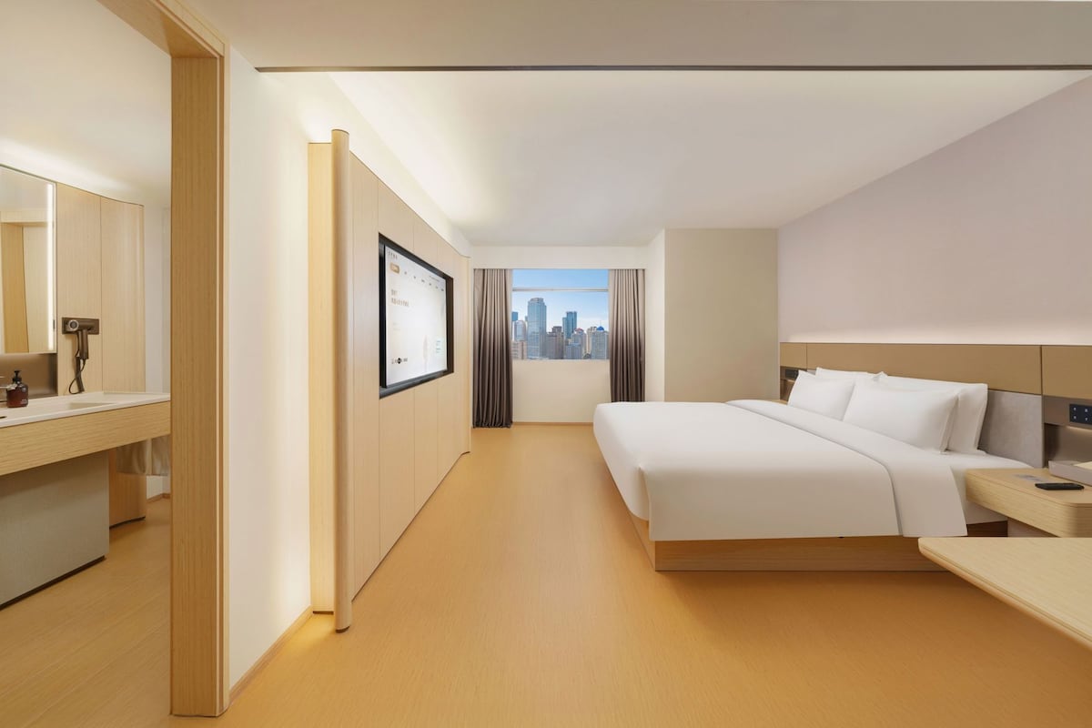 A hotel room with a bed, a bedside table, a mirror, a window with a city view, and a wall-mounted screen.