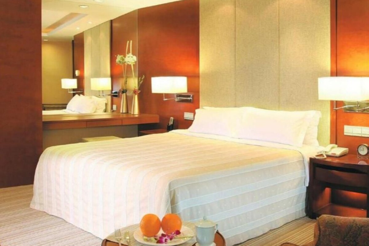 Une chambre d’hôtel avec un grand lit, des lampes de chevet, une table de nuit, un miroir et une petite table sur laquelle se trouvent une assiette de fruits et une tasse.