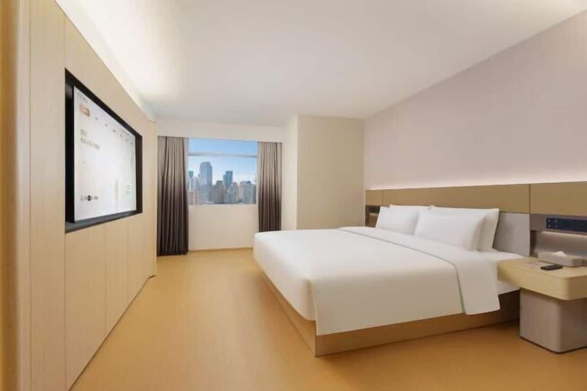 A hotel room with a bed, a nightstand, a wall-mounted TV, and a window with a cityscape view.
