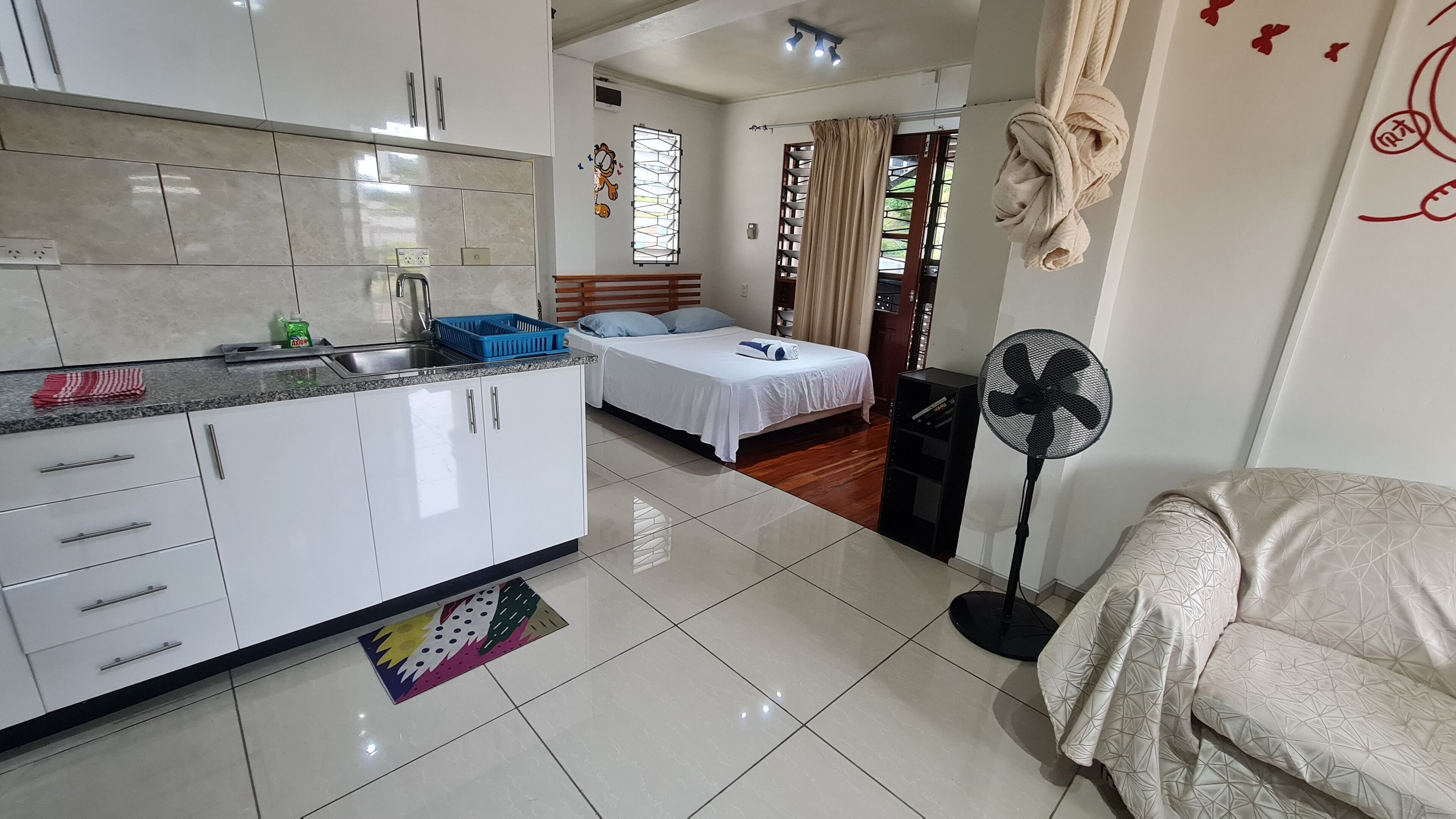 A modern hotel room with a kitchenette, a bed, a bedside table, a fan, and a window with curtains.