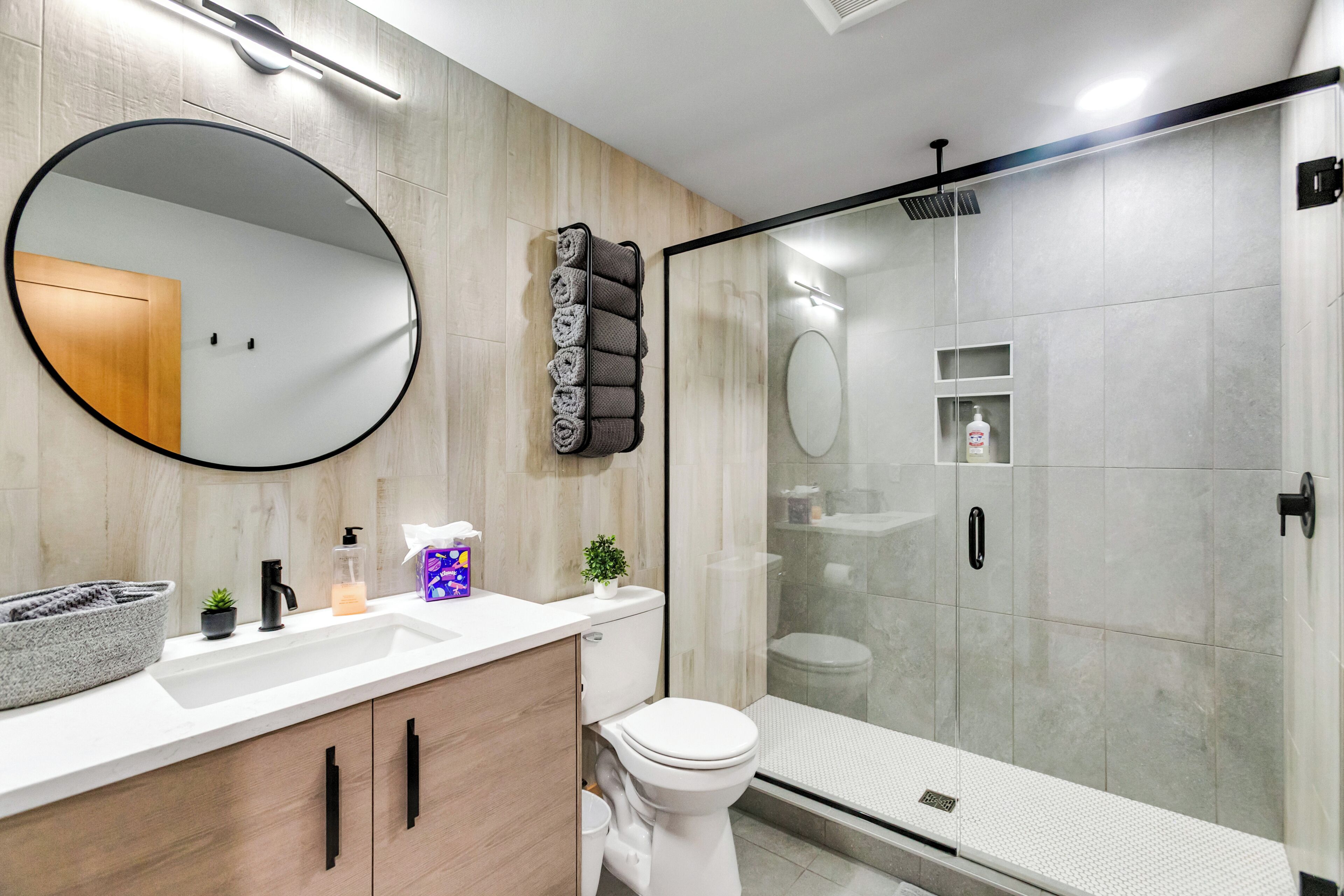 A modern bathroom with a round mirror, a toilet, a sink, a shower enclosure, and a towel rack.