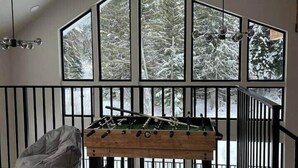 A room with a foosball table, a bean bag chair, and large windows with a snowy view. - LUXE Hyak Retreat w/SAUNA, Hot Tub, Game Room, 6BR (Snoqualmie Pass)