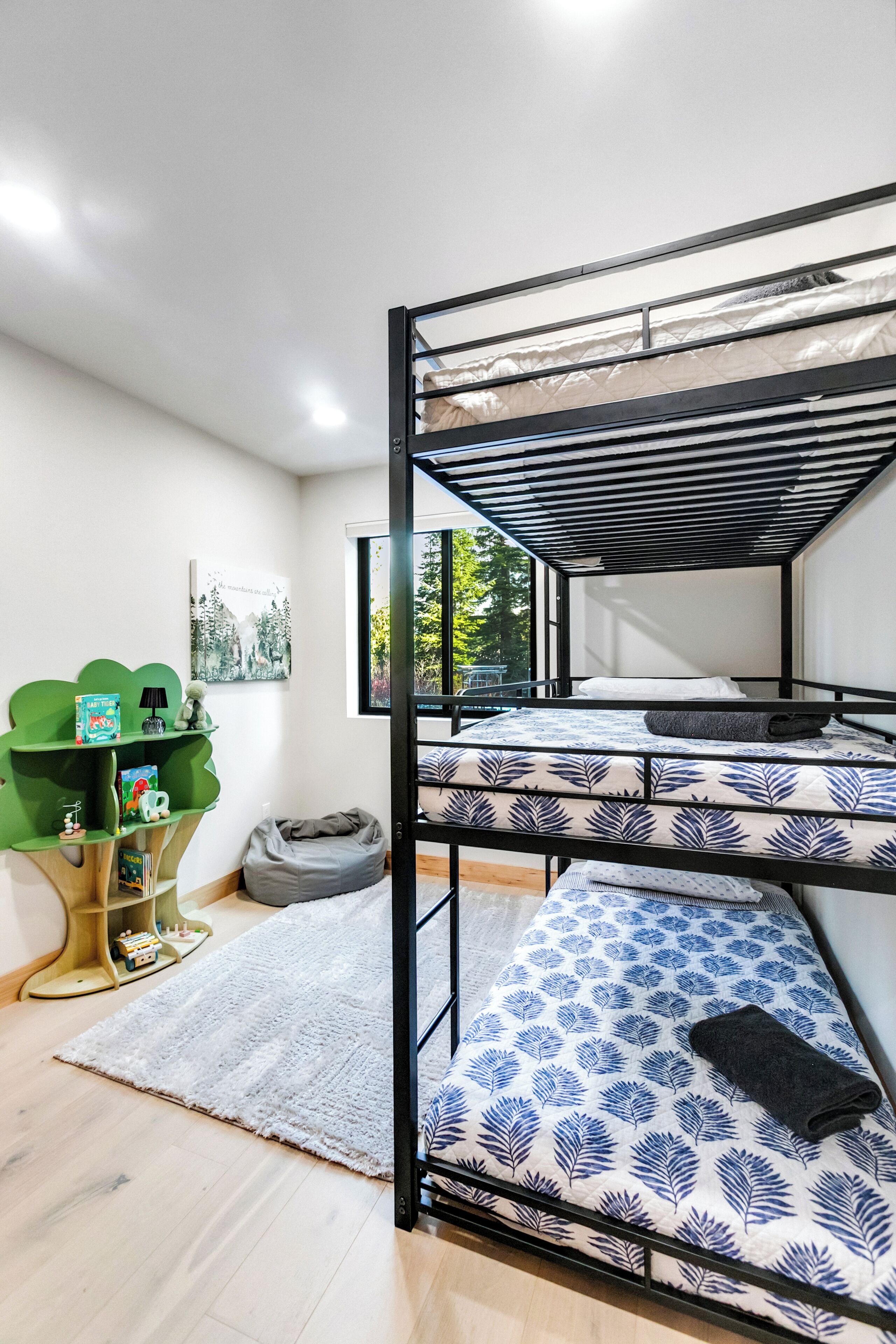 Une chambre avec un lit superposé, une étagère verte, un petit tapis et une fenêtre donnant sur des arbres.