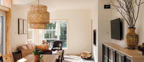 A modern dining area with a wooden table, chairs, and a hanging rattan light fixture. There is a large window providing natural light and a view of the outdoors.