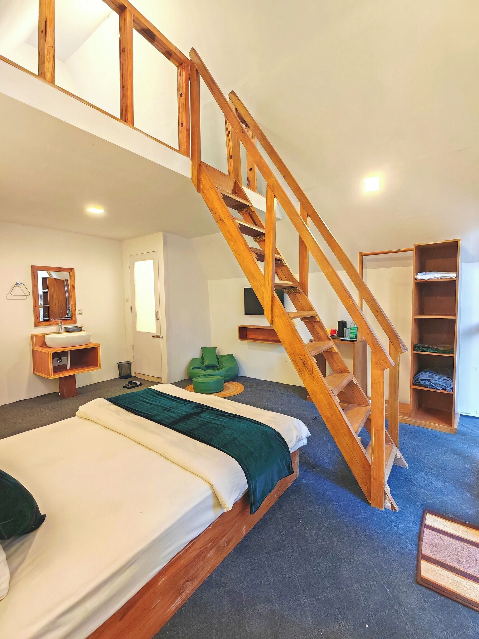 A bedroom with a wooden staircase, a bed, a bedside table, a mirror, a green bean bag chair, and a shelf with folded clothes.