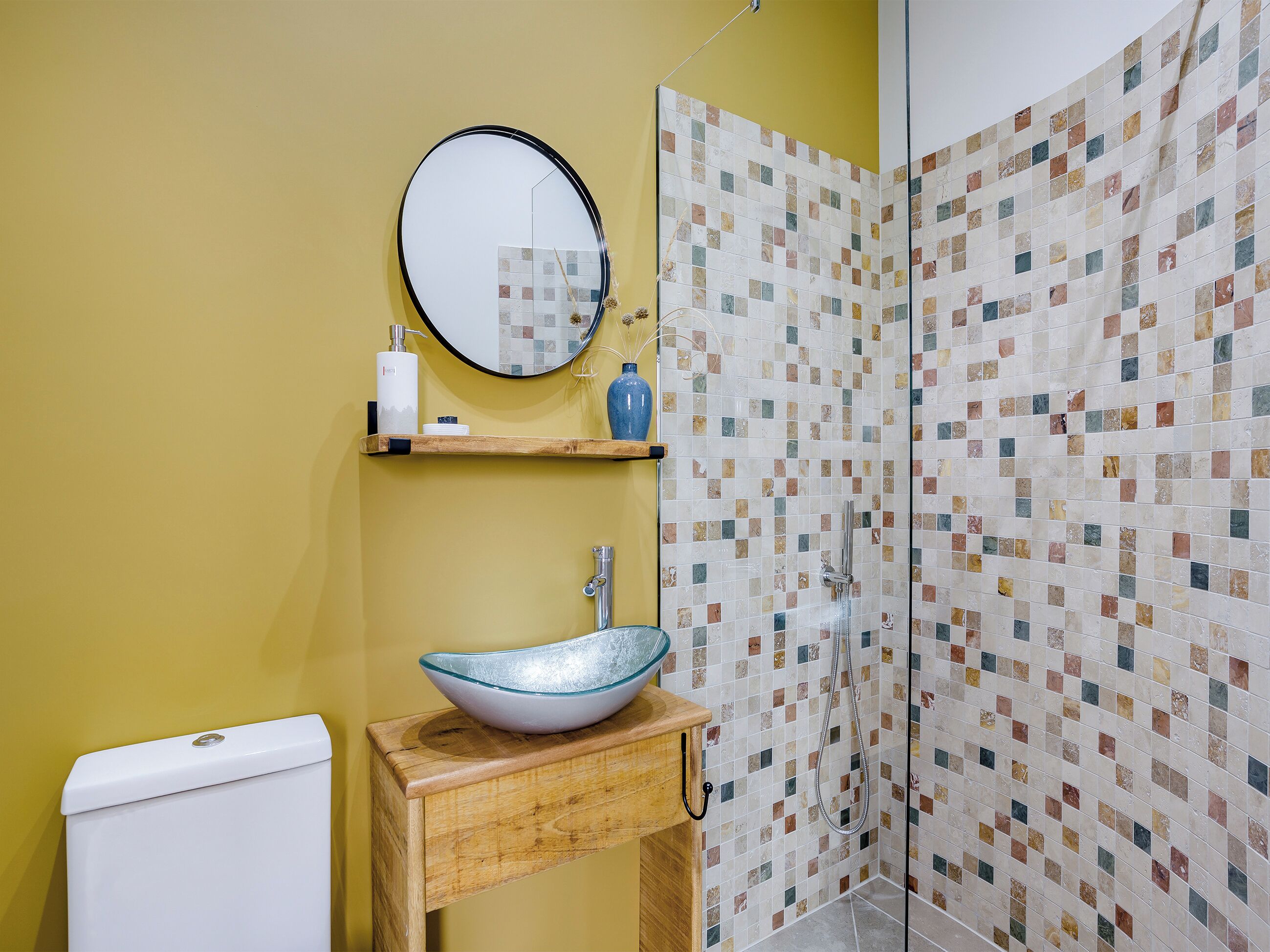 A bathroom with a toilet, a sink with a glass bowl, a mirror, and a shower with mosaic tiles.
