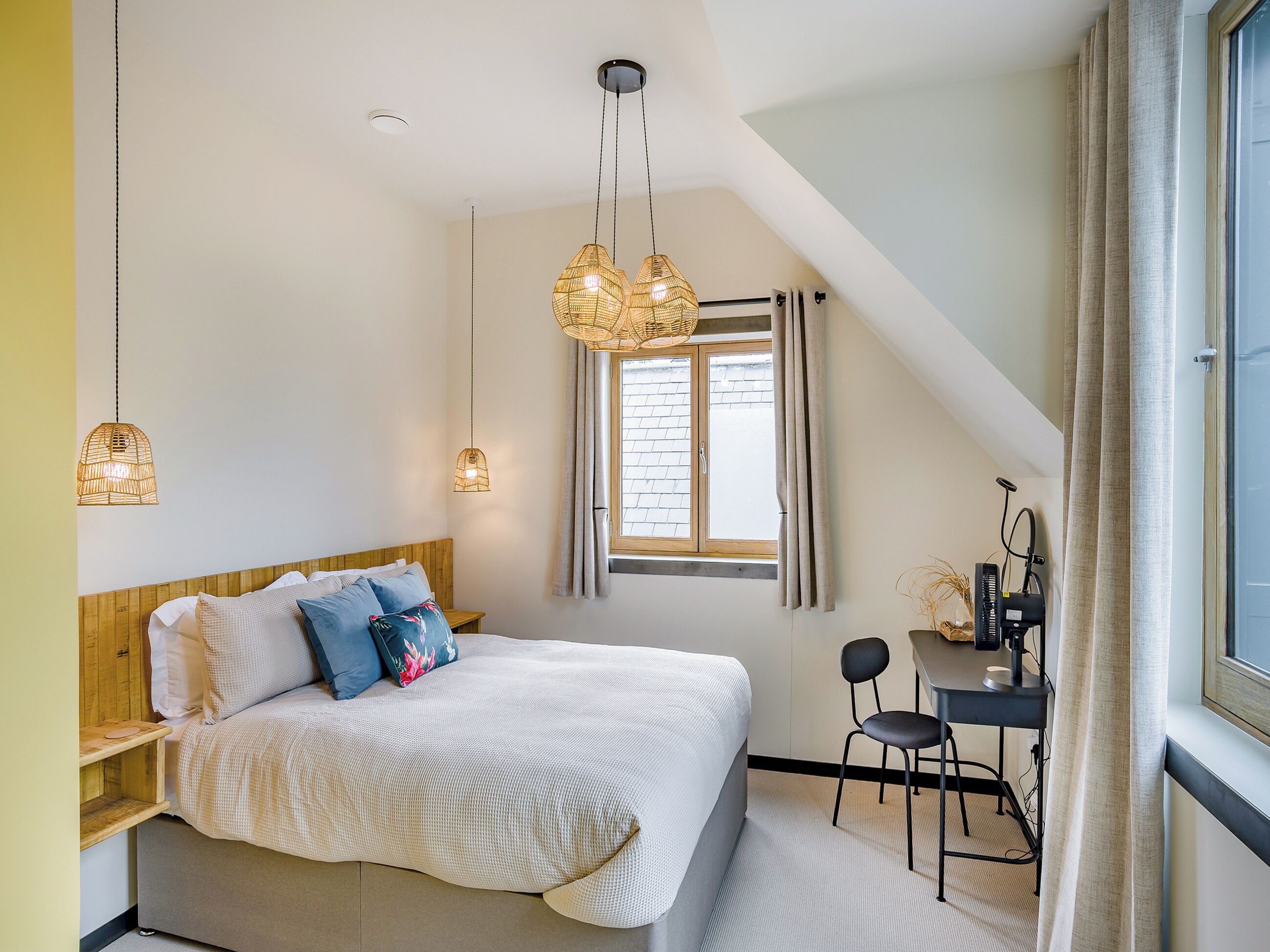 Une chambre à coucher avec un lit, deux lampes suspendues, un bureau avec une chaise et une fenêtre avec des rideaux.
