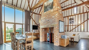 A spacious room with a large fireplace, wooden beams, a dining area, and a view through large windows.