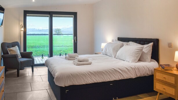 A bedroom with a large bed, a chair, a nightstand, and a view of a green field through a glass door. - Acre Luxury Lodges-Cedar Lodge (Walton Presteigne)