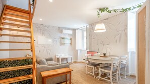 A modern living room with a wooden staircase, a grey sofa, a round dining table with white chairs, and a large window. - GuestReady - Autumn Apartment (Bairro Alto)