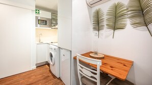 A compact kitchen with a washing machine, a microwave, and a small dining area with a wooden table and white chair. - GuestReady - Summer Studio (Bairro Alto)