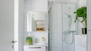 Un bany modern amb rajoles blanques, un cubicle de dutxa de vidre, un vàter blanc i un lavabo amb un mirall i una planta.
