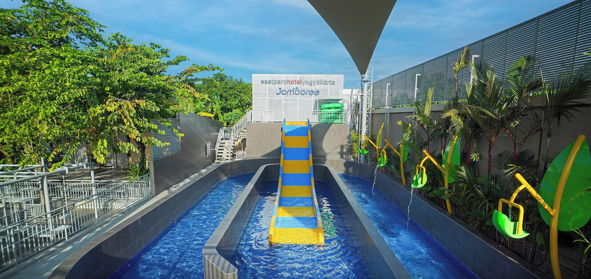 Eine Rutsche mit blau-gelbem Design, umgeben von einem Pool, Palmen und einem Schild mit der Aufschrift 'eastparchotelyogyakarta Jamboree'.