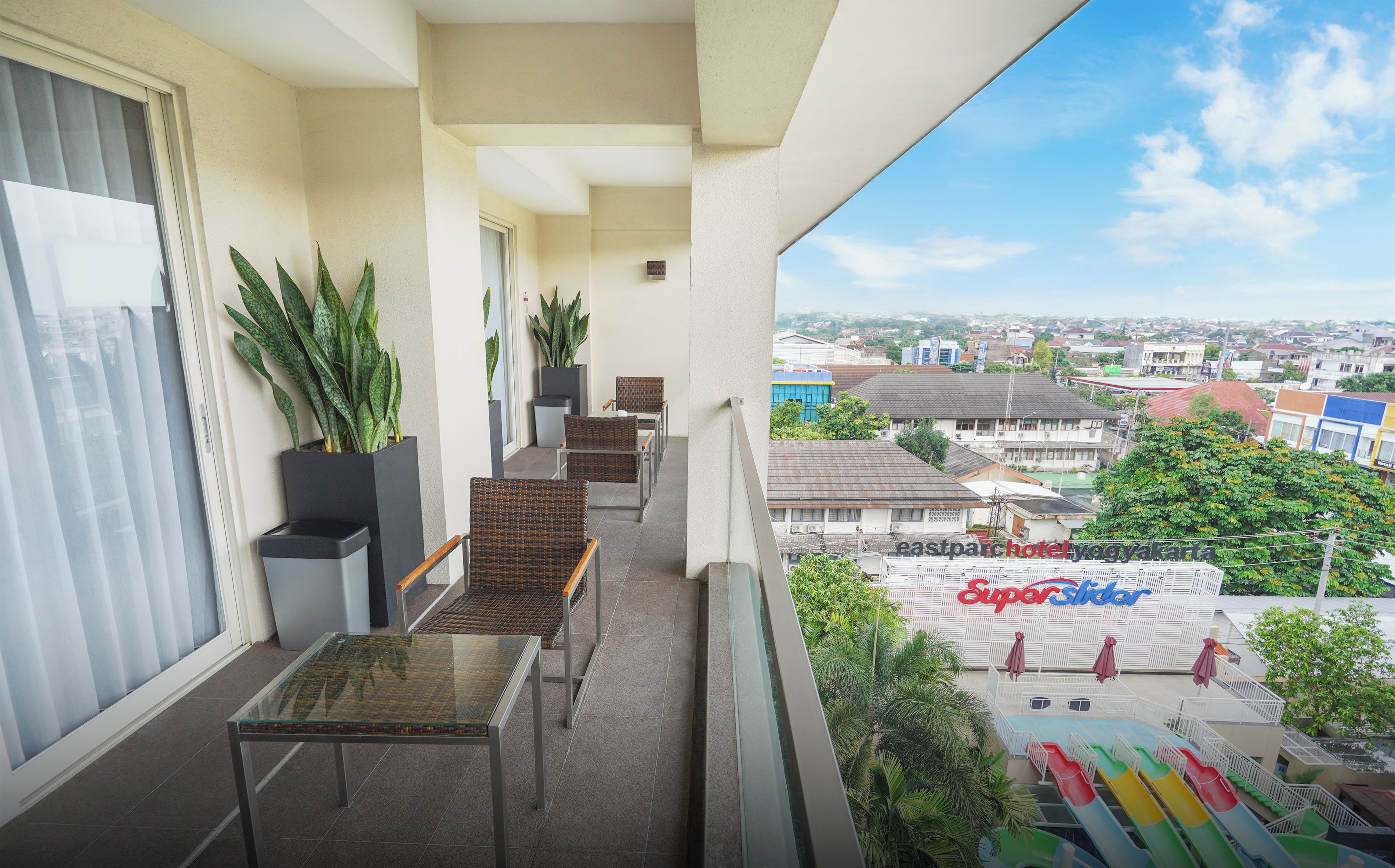 A balcony with a glass table, wicker chairs, potted plants, and a view of a water park.