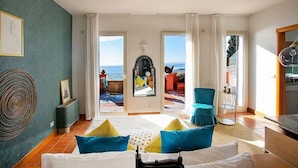 Habitación de hotel con cama, vista al mar, balcón con mobiliario exterior y zona de comedor.
