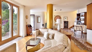 A spacious living room with a large beige sofa, a glass coffee table, and a wooden dining table. There is a large window with curtains, a fireplace, and a bookshelf. - Villa Doride Grande: An elegant and welcoming three-story residence which faces the sun and the sea, with Free WI-FI. (Taormina)