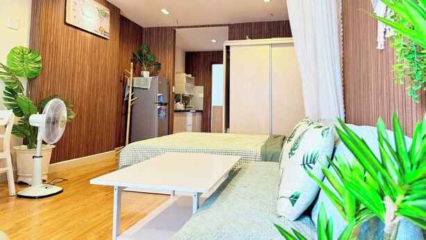 A modern room with a wooden accent wall, a white bed, a small table, a refrigerator, and a plant. - HOMESTAY 2 GIUONG DOI GAN SAN BAY TAN SON NHAT (Thành phố Hồ Chí Minh)
