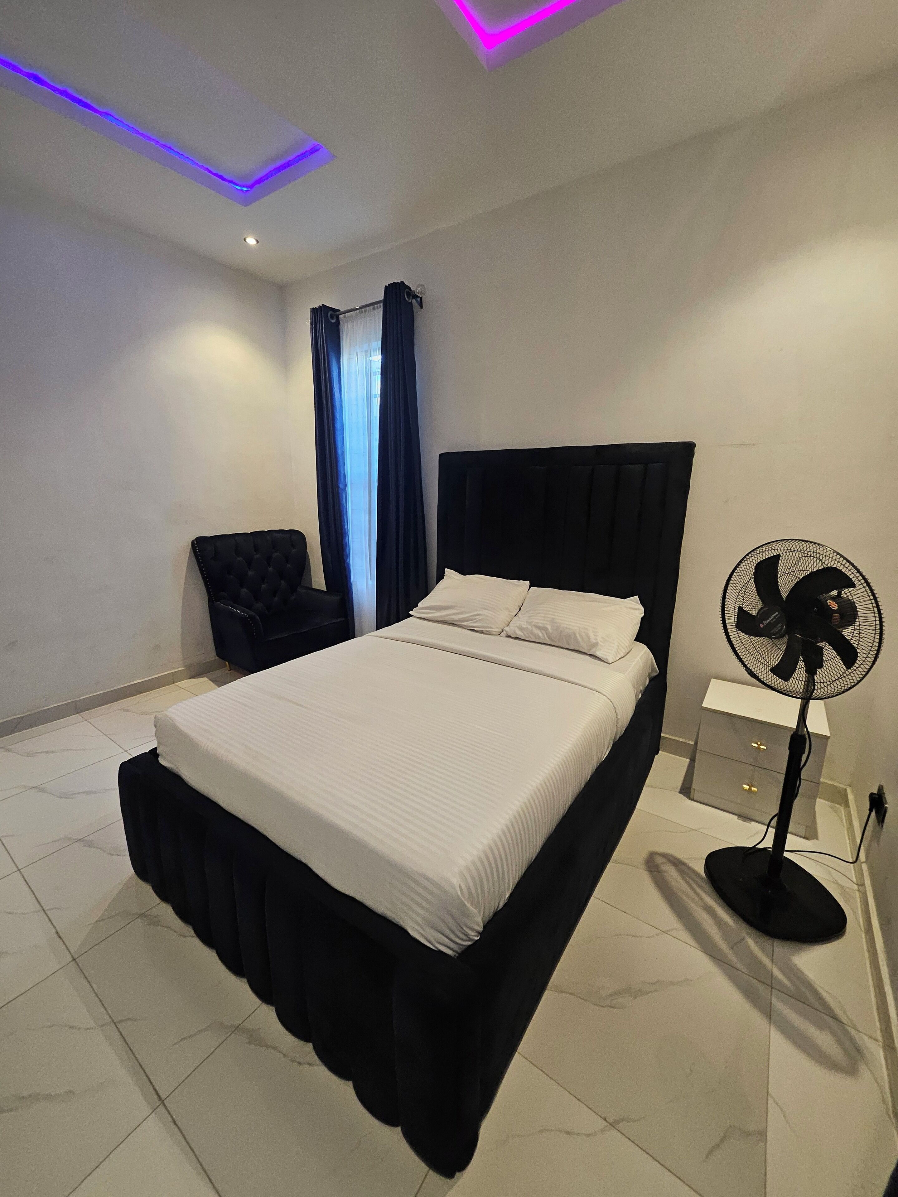A modern bedroom with a bed, a chair, a nightstand, and a standing fan.