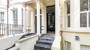 Eine Treppe führt zu einer schwarzen Tür, die von weißen Säulen gesäumt ist und einen Balkon mit schwarzer Brüstung aufweist.