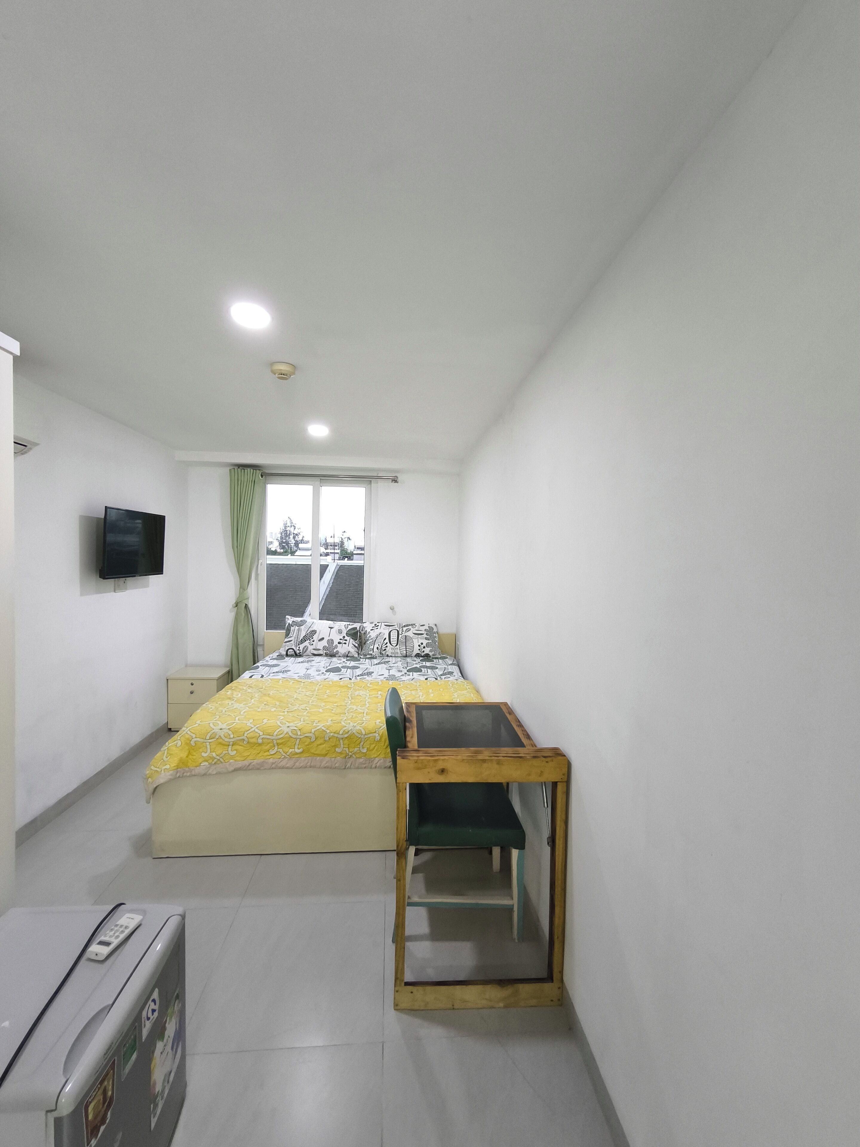 Un dormitorio con cama, un escritorio pequeño, televisor en la pared y ventana con cortinas.