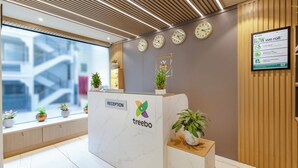 A reception desk with a marble top, a logo, and a sign indicating 'Reception'. There are clocks on the wall, potted plants, and a window with a view of buildings. - Treebo Liyan Orchid, Hsr Layout (Bengaluru)