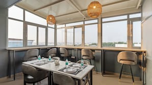 A modern dining area with a high table set for four, surrounded by chairs, large windows offering a city view, and two hanging wicker lamps. - Treebo Liyan Orchid, Hsr Layout (Bengaluru)