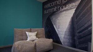 Ein Zimmer mit einer türkisfarbenen Wand, einem großen Poster mit der Aufschrift "Welcome to summit house" und einem beigen Sofa mit Kissen.