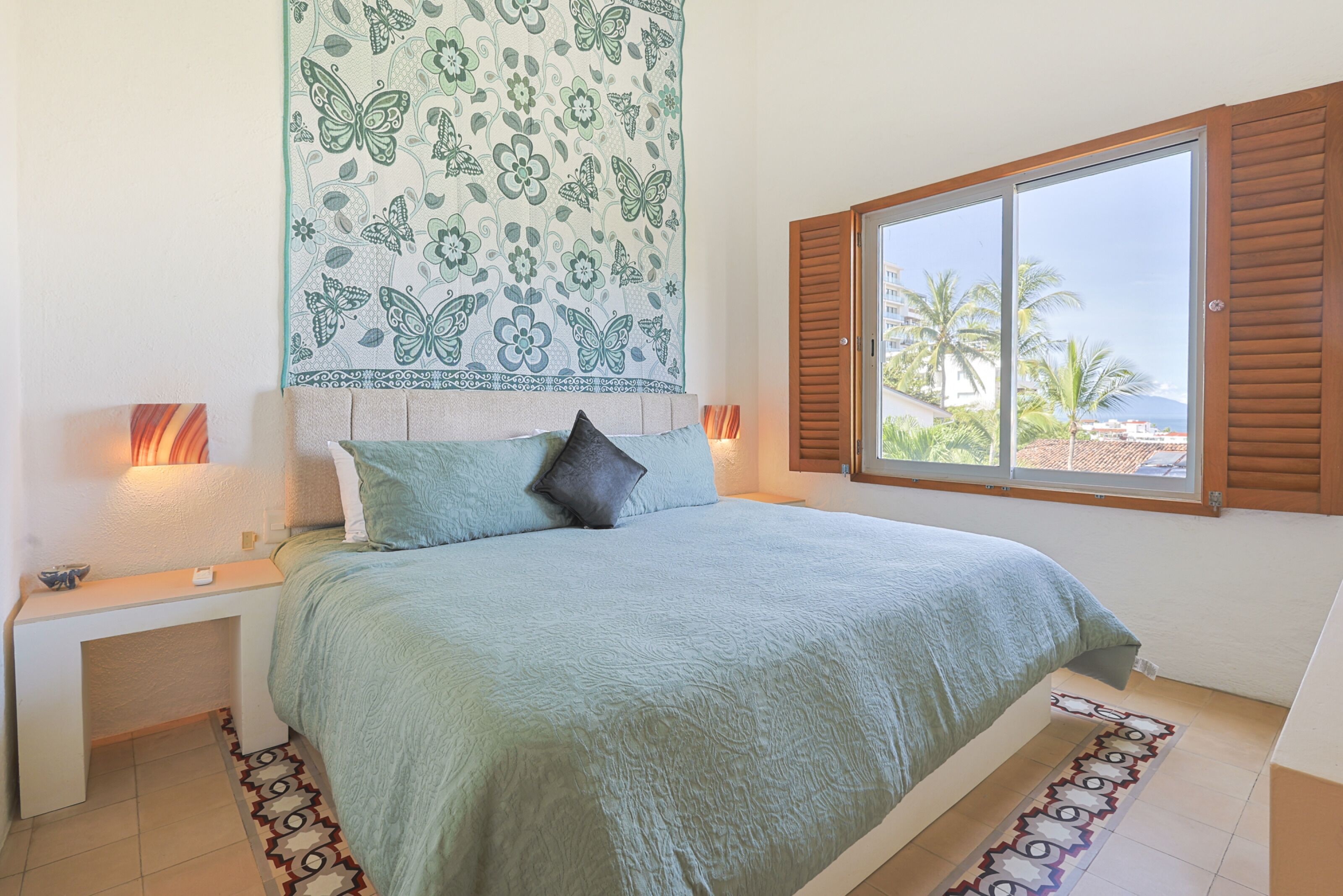 A bedroom with a bed, a nightstand, a window with shutters, and a view of palm trees and buildings.