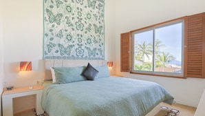 A bedroom with a bed, a nightstand, a window with shutters, and a view of palm trees and buildings. - Condo for rent in Old Town #3519 2BD Condo for rent in Old Town, Puerto vallarta (Puerto Vallarta)
