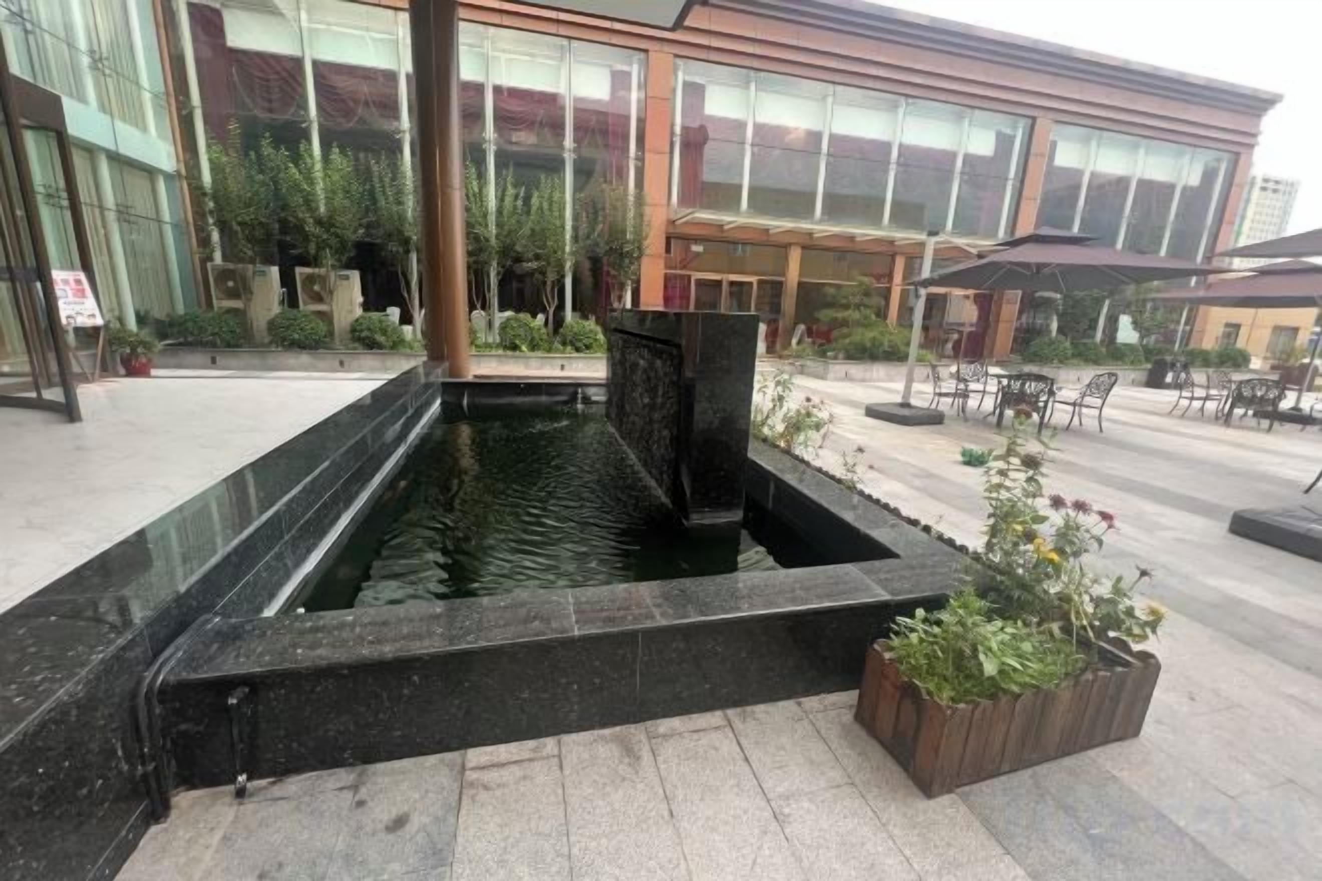 A rectangular water feature with a black border, surrounded by a paved area with potted plants.