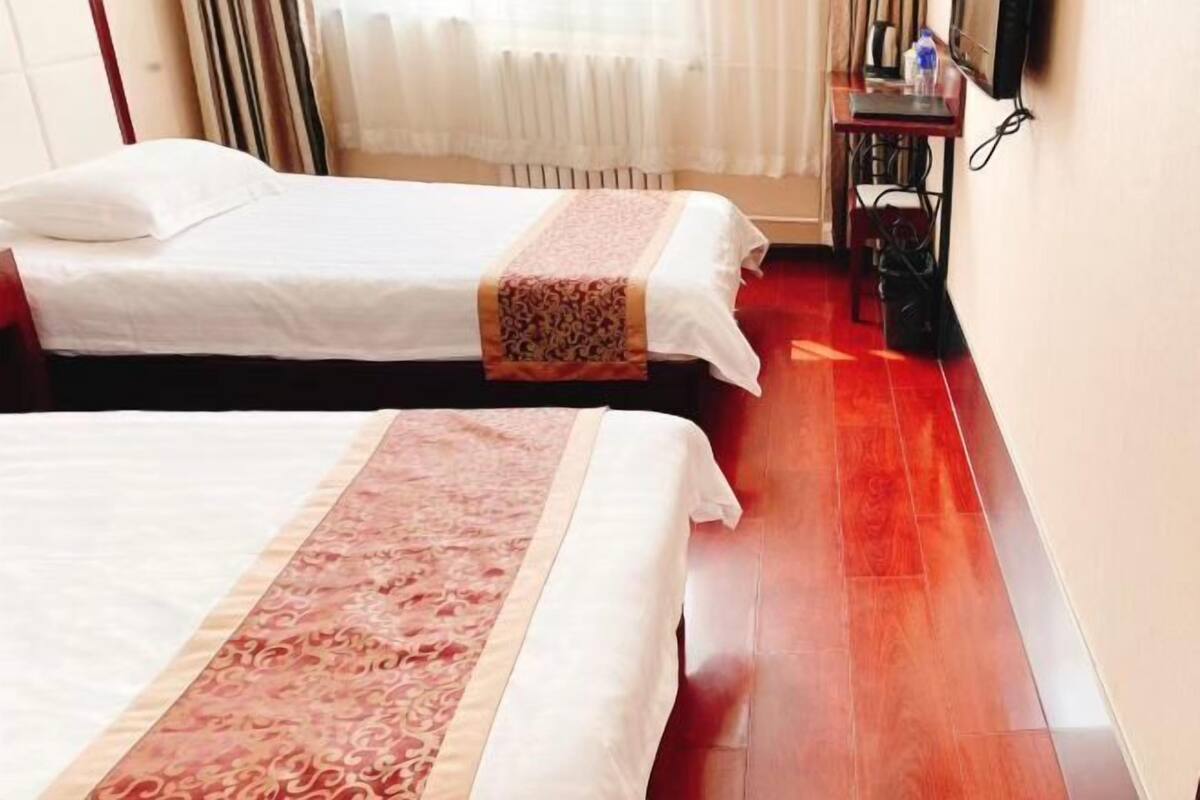 Dos camas con sábanas blancas y colchas estampadas, piso de madera, un radiador, un televisor sobre un mueble de madera y una ventana con cortinas.