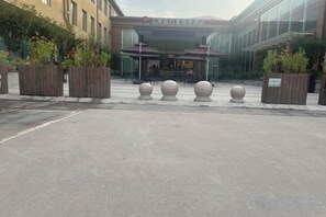 L’entrée d’un hôtel, avec une allée pavée, des jardinières en bois ornées de plantes et une façade de bâtiment au style moderne.