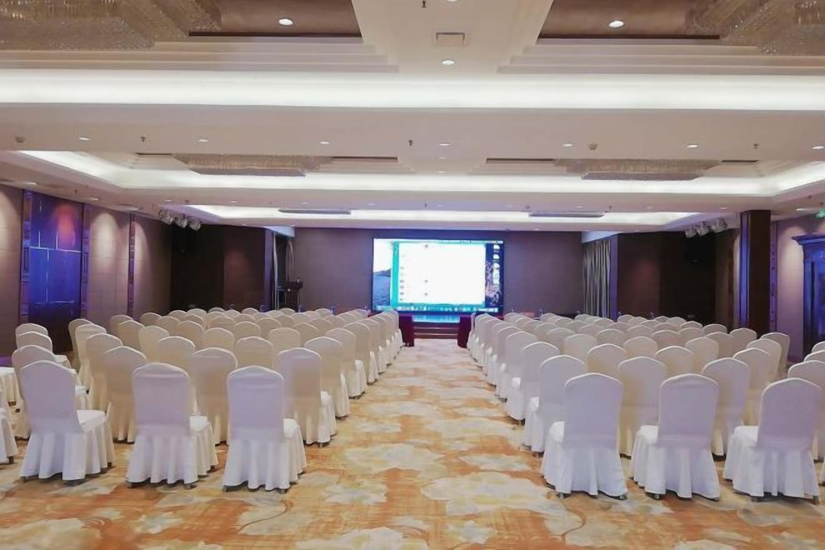 A conference room with rows of white chairs, a large screen at the front, and a carpeted floor.