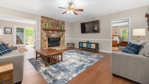 A living room with a stone fireplace, a ceiling fan, a flat-screen TV, and a large window. - Peaceful Kannapolis Lodge! Super Spacious, Fenced Yard For Pets, Near Concord Mills Mall & Speedway! (Kannapolis)