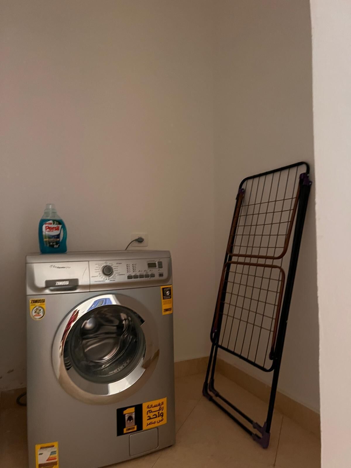 A front-loading washing machine with a detergent bottle on top, a metal drying rack, and a tiled floor.