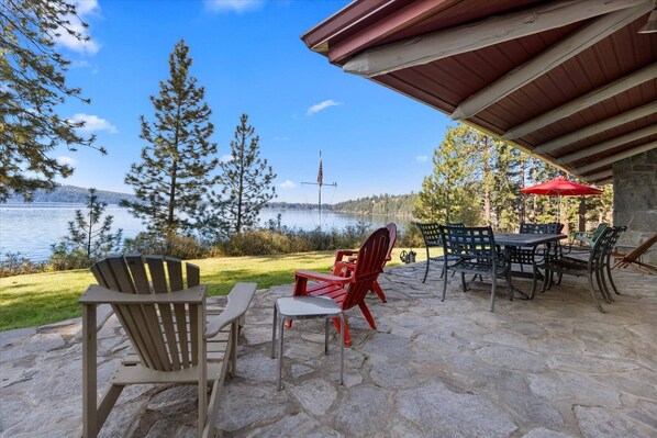 An outdoor patio with stone flooring, Adirondack chairs, a table, and a red umbrella. - Vintage Lake Retreat | Lakefront Home on Hayden Lake | Private Boat Dock & Beach (Hayden)