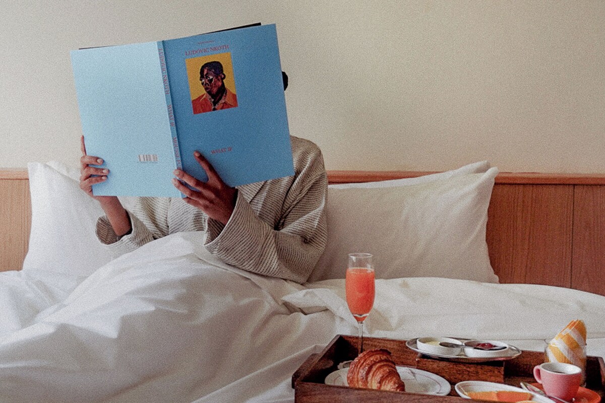 Eine Person liest im Bett ein Buch, neben ihr ein Tablett mit Frühstücksleckereien wie Croissants, Toast und einem Glas Saft.