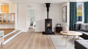 A modern living room with a wood-burning stove, a sofa, a coffee table, and a kitchen area with wooden cabinets. - 5 Star Holiday Home in Slagelse (Slagelse)