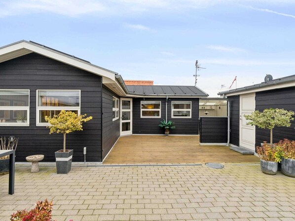 Ein schwarzes, zweistöckiges Haus mit einer Holzdachterrasse, Kübelpflanzen und einer sichtbaren Satellitenschüssel.