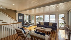 A spacious living area with a wooden floor, a sofa, chairs, and a dining table. There is a staircase with white railings leading to another level. - Pine Creek Beach House (Fairfield)
