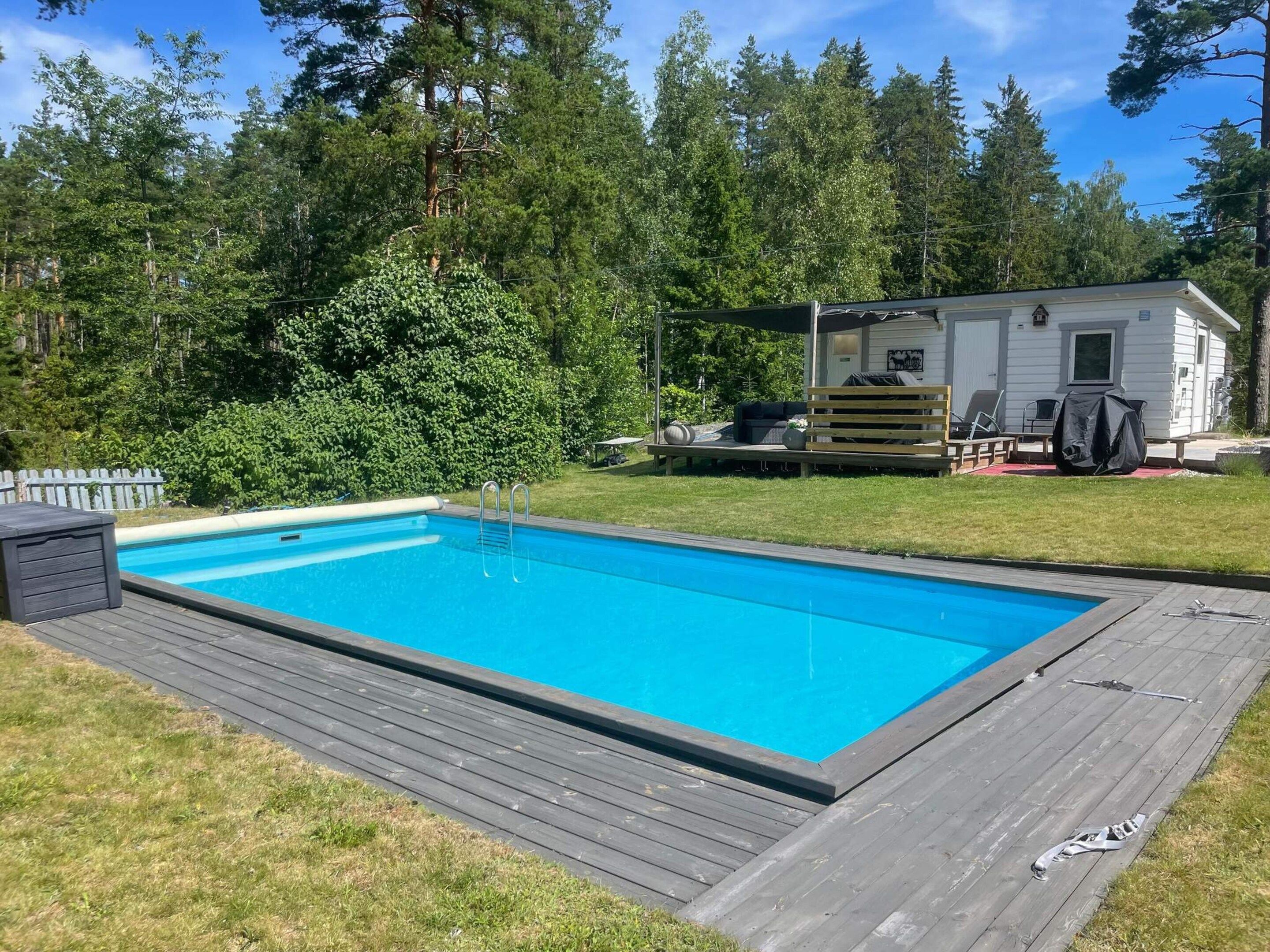 A rectangular swimming pool with a wooden deck, surrounded by a white mobile home with outdoor furniture.