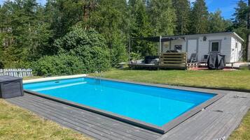 A rectangular swimming pool with a wooden deck, surrounded by a white mobile home with outdoor furniture.