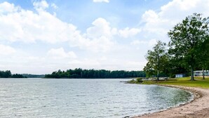 Blick auf einen See mit Sandstrand, einer Anlegestelle und einer Reihe von Hütten im Hintergrund.