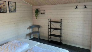 Una habitación con paredes de madera, una cama con ropa blanca, una silla gris, un estante negro y dos cuadros enmarcados de mariposas.