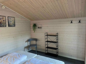 Une piÚce aux murs en bois, un lit avec une literie blanche, une chaise grise, une étagÚre noire et deux tableaux représentant des papillons, encadrés.