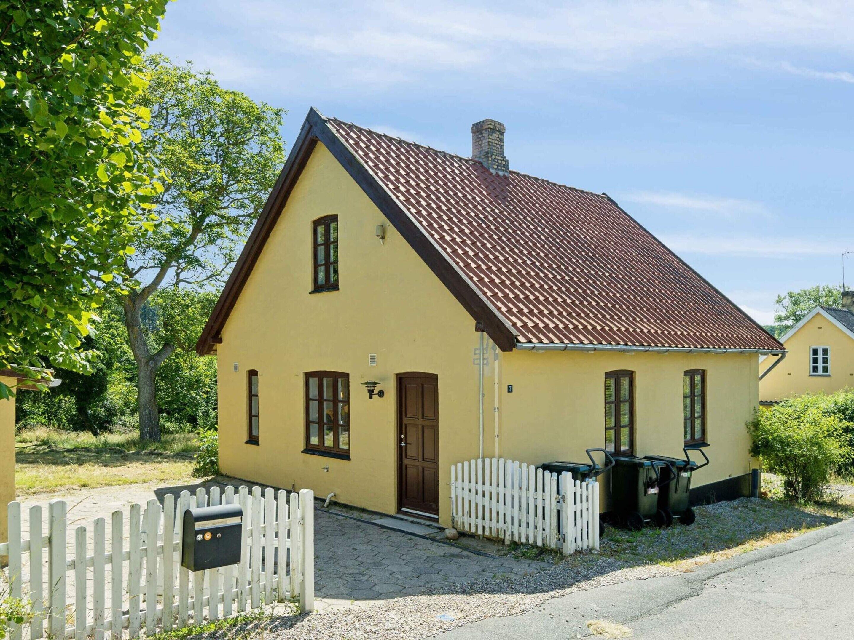 A single-story yellow house with a brown tiled roof, a white picket fence, and a small wheelbarrow near the entrance.