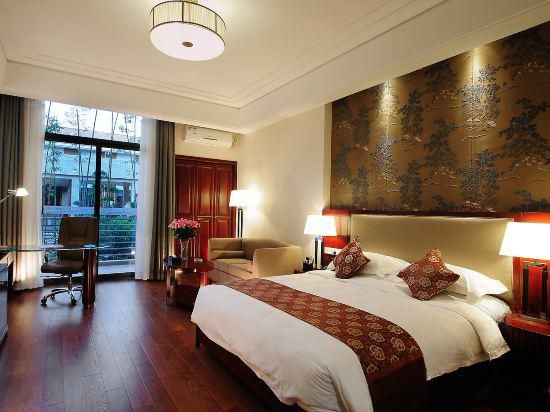 A hotel room with a large bed, two bedside lamps, a desk with a chair, a window with curtains, and a large decorative wall piece.