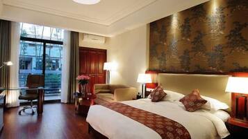 A hotel room with a large bed, two bedside lamps, a desk with a chair, a window with curtains, and a large decorative wall piece.