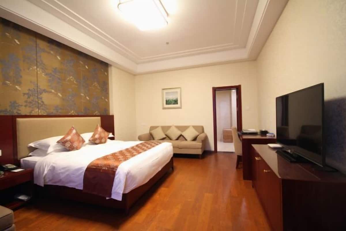 A hotel room with a bed, a sofa, a desk with a TV, and a painting on the wall.