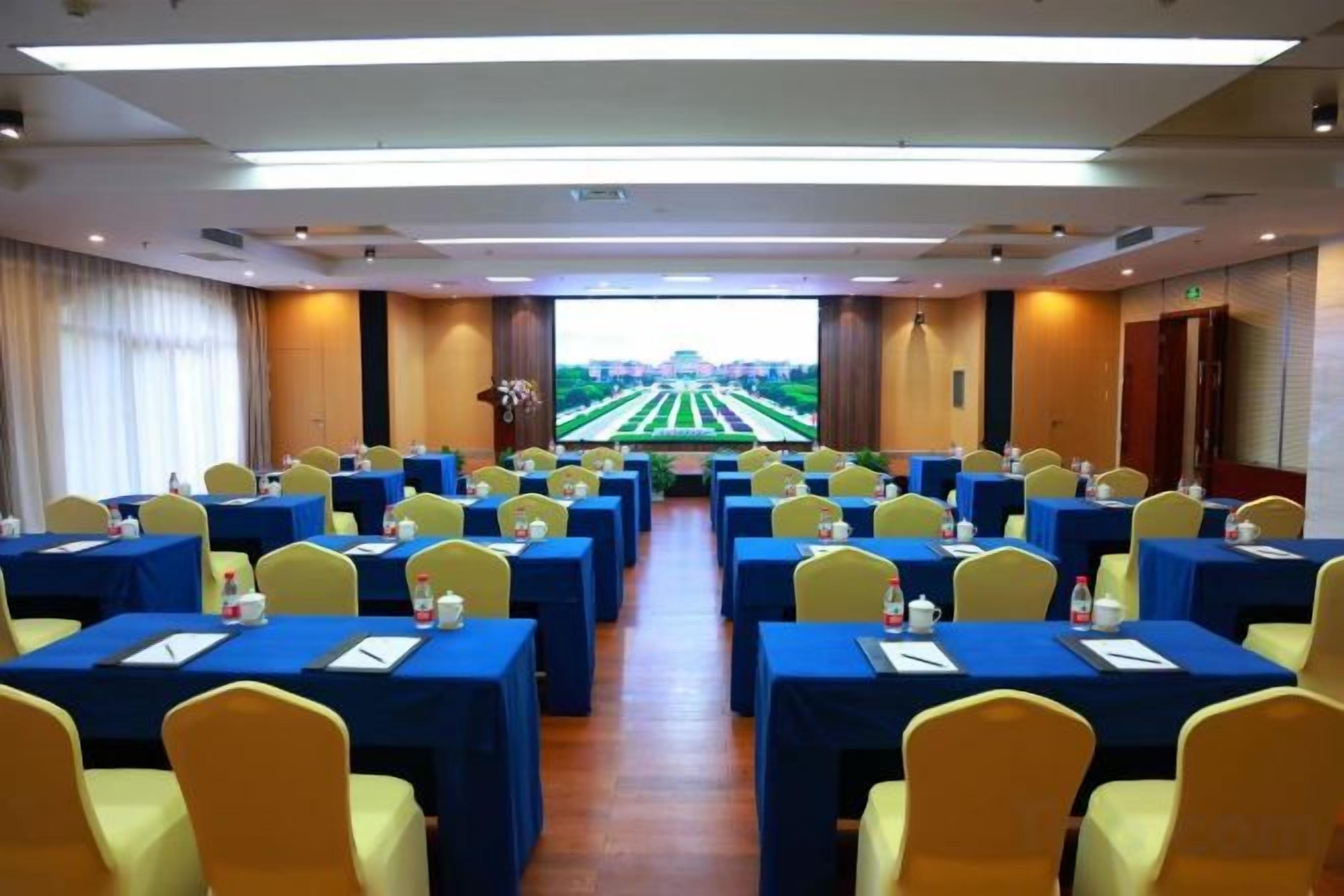 A conference room with rows of tables and chairs, a large screen at the front, and a view of a green field through the windows.
